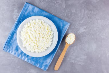 Gri çimento kaplamalı beyaz tabakta süzme peynir. Süt ürünü ve tahta kaşık. Mavi peçeteye bir tabak süzme peynir. Üst Manzara. Boşluğu kopyala