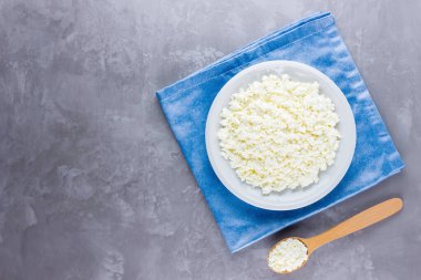 Gri çimento kaplamalı beyaz tabakta süzme peynir. Süt ürünü ve tahta kaşık. Mavi peçeteye bir tabak süzme peynir. Üst Manzara. Boşluğu kopyala