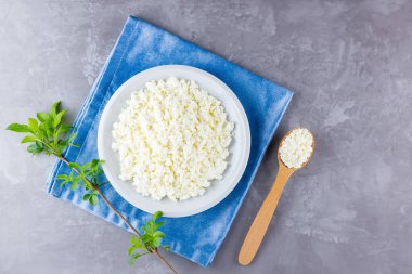 Gri arka planda beyaz tabakta süzme peynir. Yumuşak peynir ve çimentoda ahşap kaşık. Mavi peçetede yeşil yapraklı süzme peynir ve dal. Üst Manzara. Boşluğu kopyala