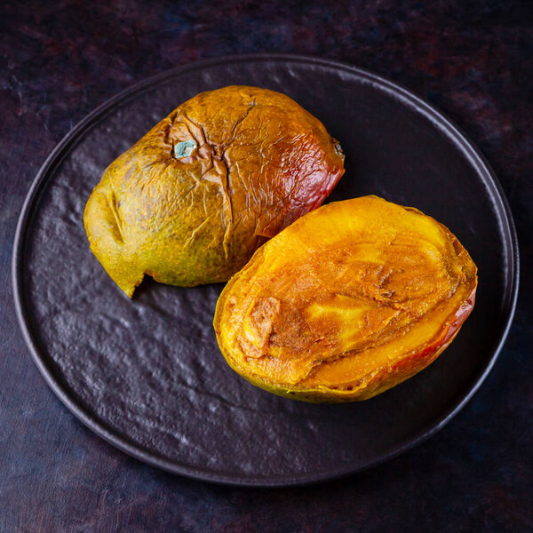 Уродливое манго. Испорченный манго на темном фоне. Гнилое манго на черной тарелке. Вид сверху. Копирование пространства
