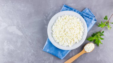 Gri arka planda beyaz tabakta süzme peynir. Yumuşak peynir ve çimentoda ahşap kaşık. Mavi peçetede yeşil yapraklı süzme peynir ve dal. Üst Manzara. Boşluğu kopyala