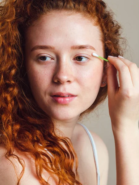 The concept of removing makeup from the face. Attractive girl with long red curly hair corrects cosmetics on the skin. No retouching. Natural beauty. Close-up shot in studio