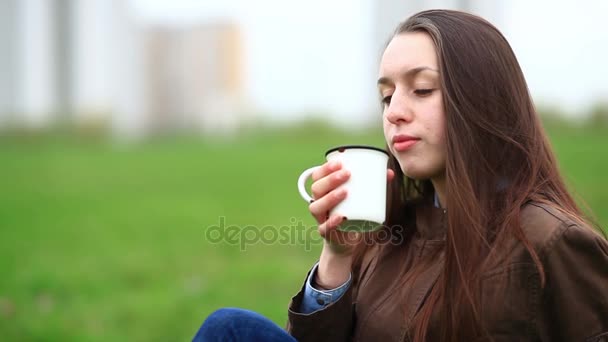 Młoda dziewczyna z płynącej włosów jest picie herbaty z białym kubku zielonego parku na zewnątrz. — Wideo stockowe