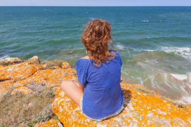 Genç bir kadın deniz kenarındaki bir kayanın üzerinde oturuyor ve rüyasında ufka bakıyor.