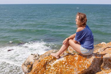 Genç bir kadın deniz kenarındaki bir kayanın üzerinde oturuyor ve rüyasında ufka bakıyor.