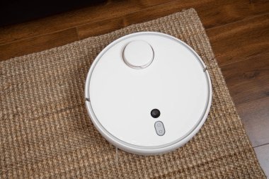 Beyaz robotik süpürge, oturma odasının iç tarafındaki plastik zeminde toz temizliyor. Akıllı elektronik temizlik teknolojisi.
