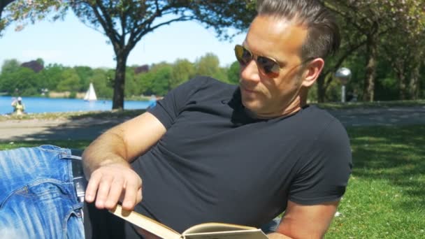Zomer in de stad - een man op het grasveld in het park ontspant en leest een boek — Stockvideo