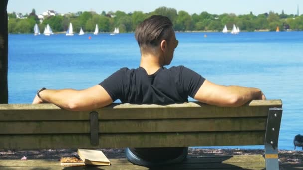 Un hombre disfruta de su tiempo libre en un banco frente a un hermoso lago — Vídeo de stock