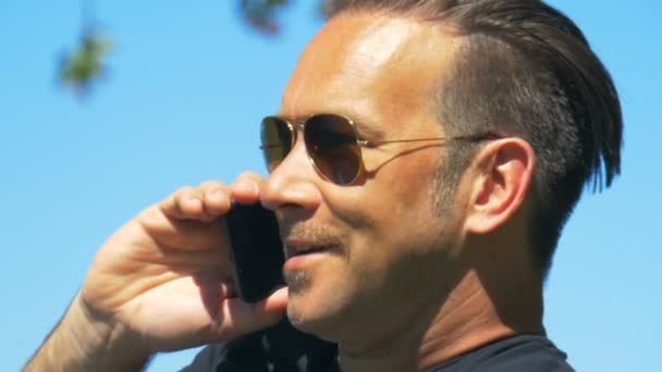 Um homem alegre de óculos de sol fala ao telefone e sorri — Vídeo de Stock