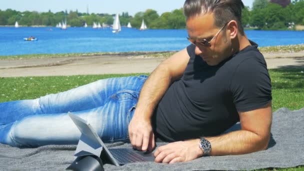 Um homem relaxa ao ar livre com bom tempo e surfa no laptop — Vídeo de Stock