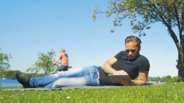 A hétvégén a városban - egy férfi ellazítja a füvön, a parkban, és elolvassa a könyvet — Stock videók