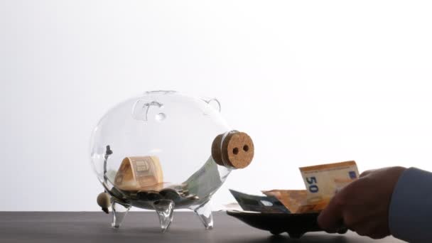 Gondoskodik a saját pénzéről - egy férfi egy bankjegyekkel teli tálat tesz egy üvegmalac bank elé, és néhány érmét is dobál bele. — Stock videók