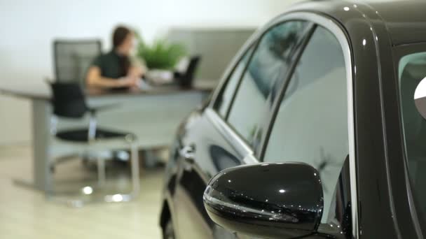 Trabalha na sala de exposições. Gerente de vendas na concessionária trabalha em uma mesa. Em primeiro plano do carro, o Gestor de imagens está desfocado . — Vídeo de Stock