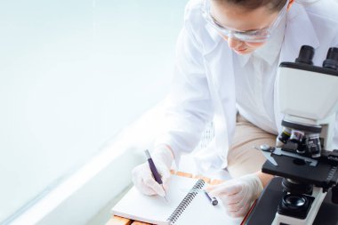 Bilim adamı doktor, hastane tıp laboratuarında mikroskopla iş günü üzerine not alıyor ya da not yazıyor.