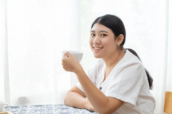 cute Asian girl teen sitting at home hand hold white cup mug and smile for diet drinking herbal tea or healthy drink.