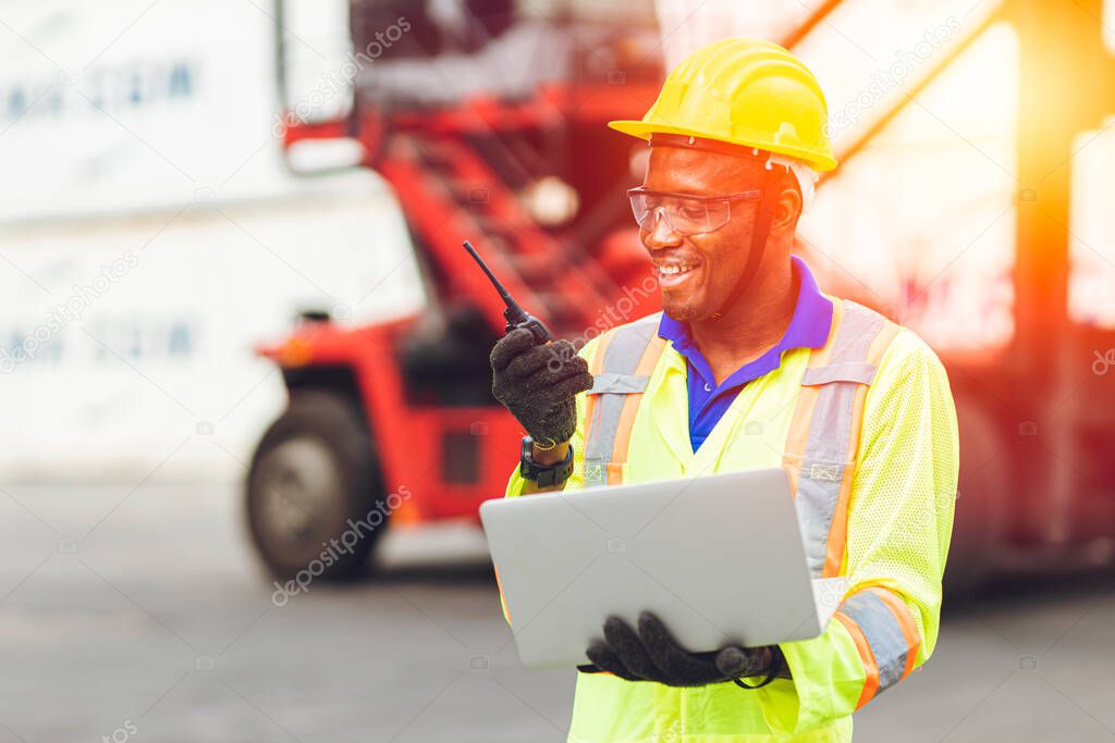 Black African happy worker working in logistic communication using radio and laptop to control loading containers at port cargo to trucks for export and import goods.