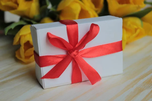 Gift in a white box with a red ribbon, on a background of yellow tulips .