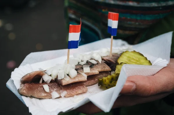 Una mano sostiene un tradicional bocadillo holandés de arenque con pepinillos y cebollas . — Foto de Stock