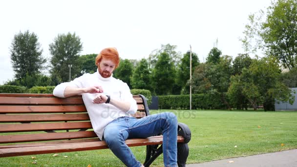 Pomocí hodinek Smartwatch, muž sedí v parku na lavičce, červené vlasy a vousy — Stock video