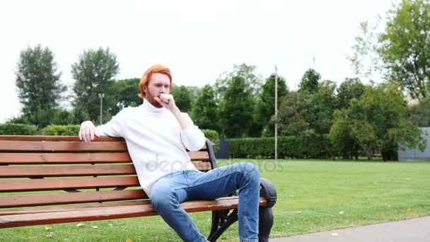 Больной кашляет, сидит в парке, рыжие волосы и борода — стоковое видео