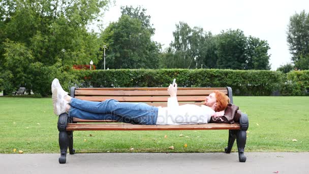 Człowiek leżący na ławce w parku i przy użyciu smartfona, czerwone włosy — Wideo stockowe