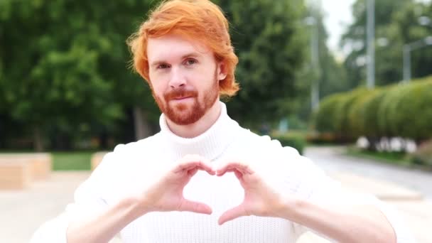 Знак сердца человека с рыжими волосами, на открытом воздухе — стоковое видео