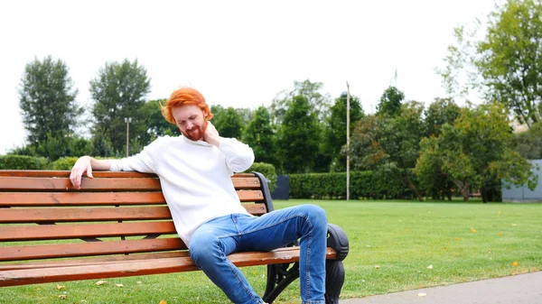 Trött Man med nacksmärta, rött skägg och hår, sitter i parken på bänk — Stockfoto