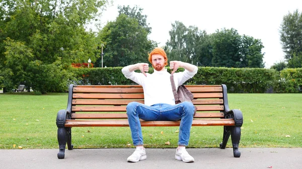 Båda händerna tummen ner av mannen som sitter på bänken i parken, röda hår och skägg — Stockfoto