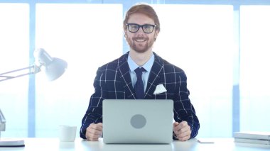 Kızıl saçlı işadamı kamera dizüstü bilgisayarda çalışırken doğru gülümseyen