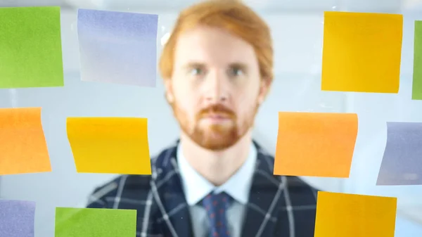 Redhead člověka dívá skrze Sticky Notes připojené na skle v úřadu — Stock fotografie