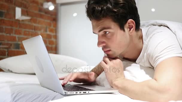 Молодой человек в постели работает над ноутбуком и реагирует на успех — стоковое видео