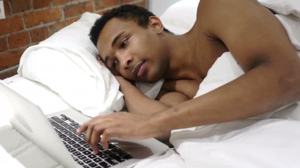 Afrykański mężczyzna pracujący na laptopie, leżąc w łóżku dla odpoczynku — Wideo stockowe