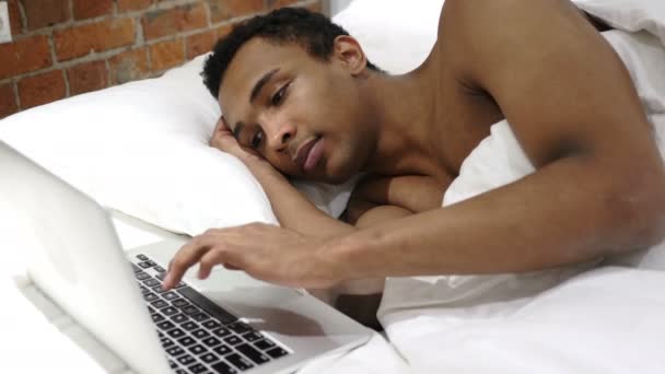 Hombre africano escribiendo correo electrónico en el ordenador portátil, acostado en la cama para el descanso — Vídeos de Stock
