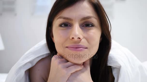 Mujer acostada en la cama sonriendo y mirando a la cámara, de cerca — Vídeos de Stock