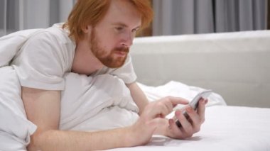 Kaydırma ve Smartphone üzerinde tarama yatakta yatan kızıl saçlı sakallı adam