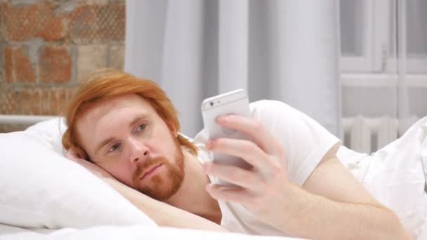 Pelirroja barba hombre acostado en la cama y navegar en el teléfono inteligente por la noche — Vídeo de stock