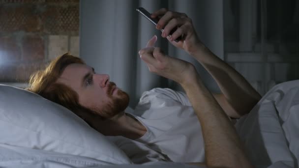 Mann mit rothaarigem Bart surft E-Mails und Nachrichten am Telefon im Bett — Stockvideo