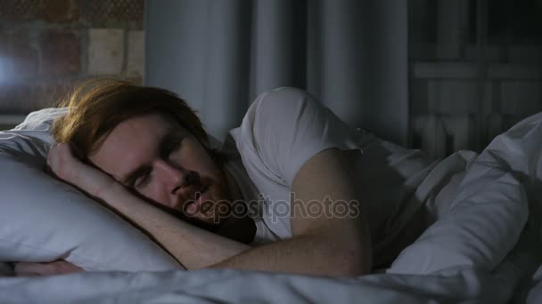 Рыжеволосый бородач лежит в постели и спит в темной комнате ночью — стоковое видео
