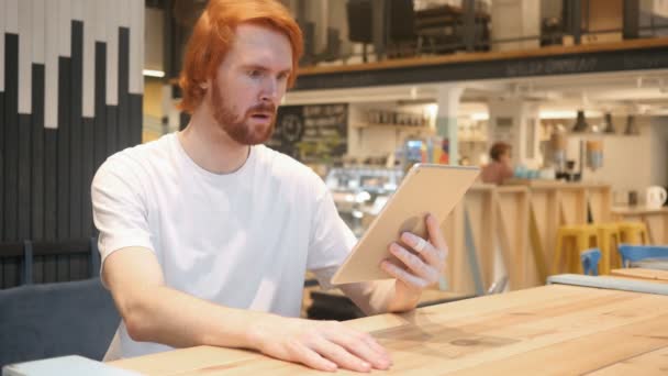 Шокированный, удивляющийся рыжеволосый человек, использующий планшетный компьютер в кафе — стоковое видео
