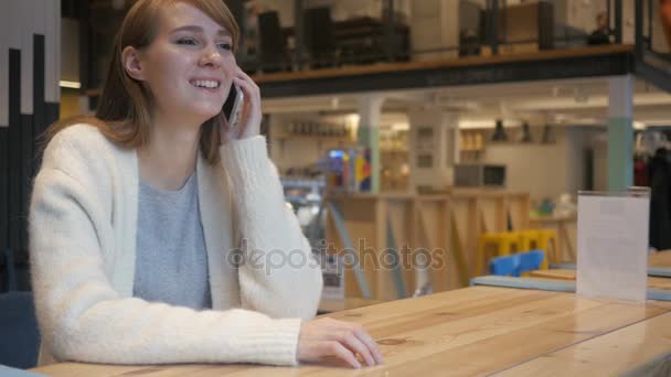 Молодая женщина разговаривает по телефону, сидя в кафе — стоковое видео