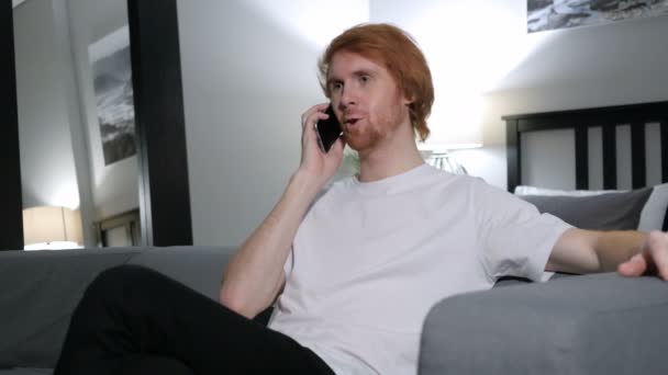 Ryšavý muž mluví o telefonu, sedící na gauči v ložnici — Stock video