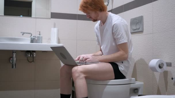 Rotschopf tippt auf Laptop, sitzt auf Toilettenkommode — Stockvideo