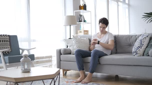 Młody człowiek za pomocą Smartwatch jednocześnie relaksująca na kanapie — Wideo stockowe