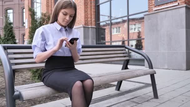 Femme utilisant un téléphone intelligent, naviguant en étant assise à l'extérieur de l'immeuble de bureaux — Video