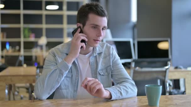 Веселый молодой дизайнер разговаривает по смартфону в офисе — стоковое видео