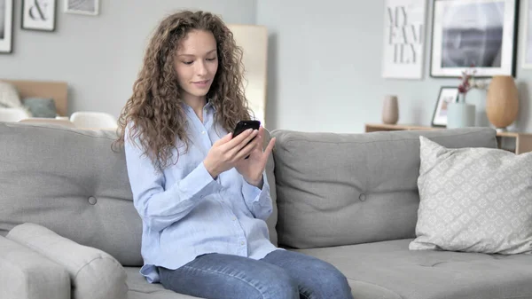 Jonge krullend haar vrouw het gebruik van smartphone terwijl ontspannen op de Bank — Stockfoto