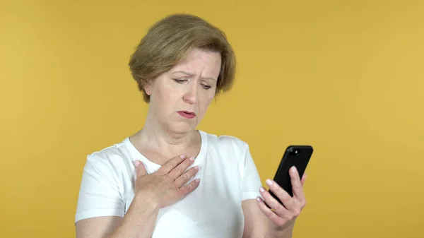 Стара жінка реагує на втрату та використання смартфона ізольовано на жовтому тлі — стокове фото