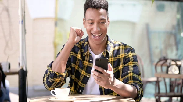Young African Man Celebrating Success on Smartphone, Outdoor Cafe