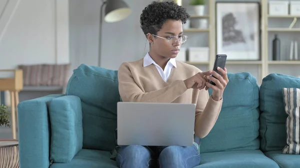 La giovane ragazza africana che lavora con telefono e laptop sul divano — Foto Stock