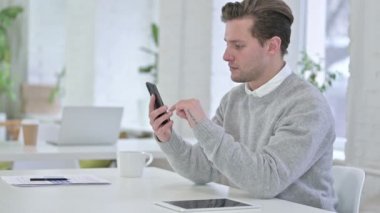Ofiste akıllı telefon kullanan neşeli yaratıcı genç adam.